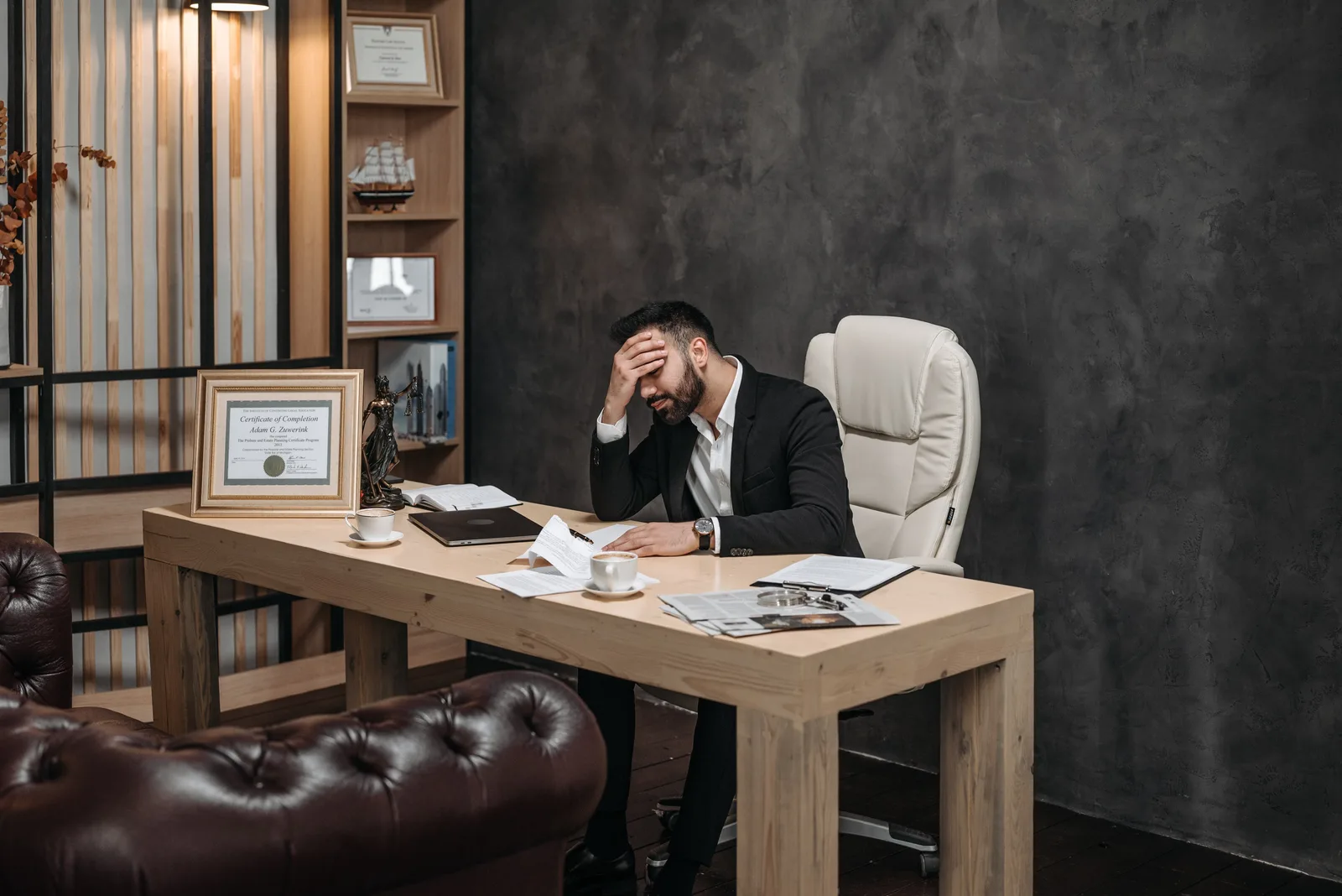 Businessman in a black suit looking stressed at an office desk, holding his head with papers scattered around.